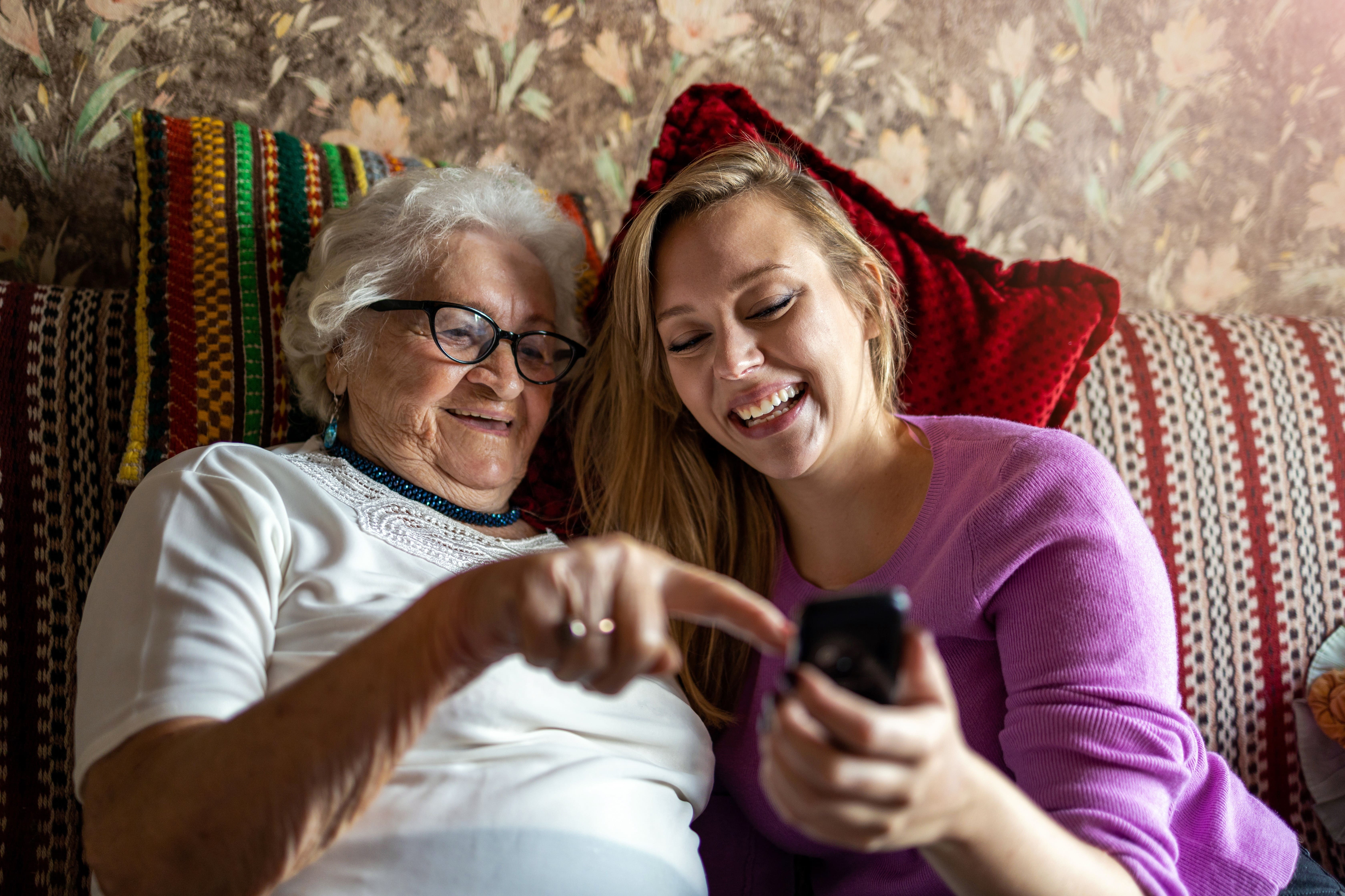 An elderly lady at her home with her female carer. They are both looking at a phone and smiling