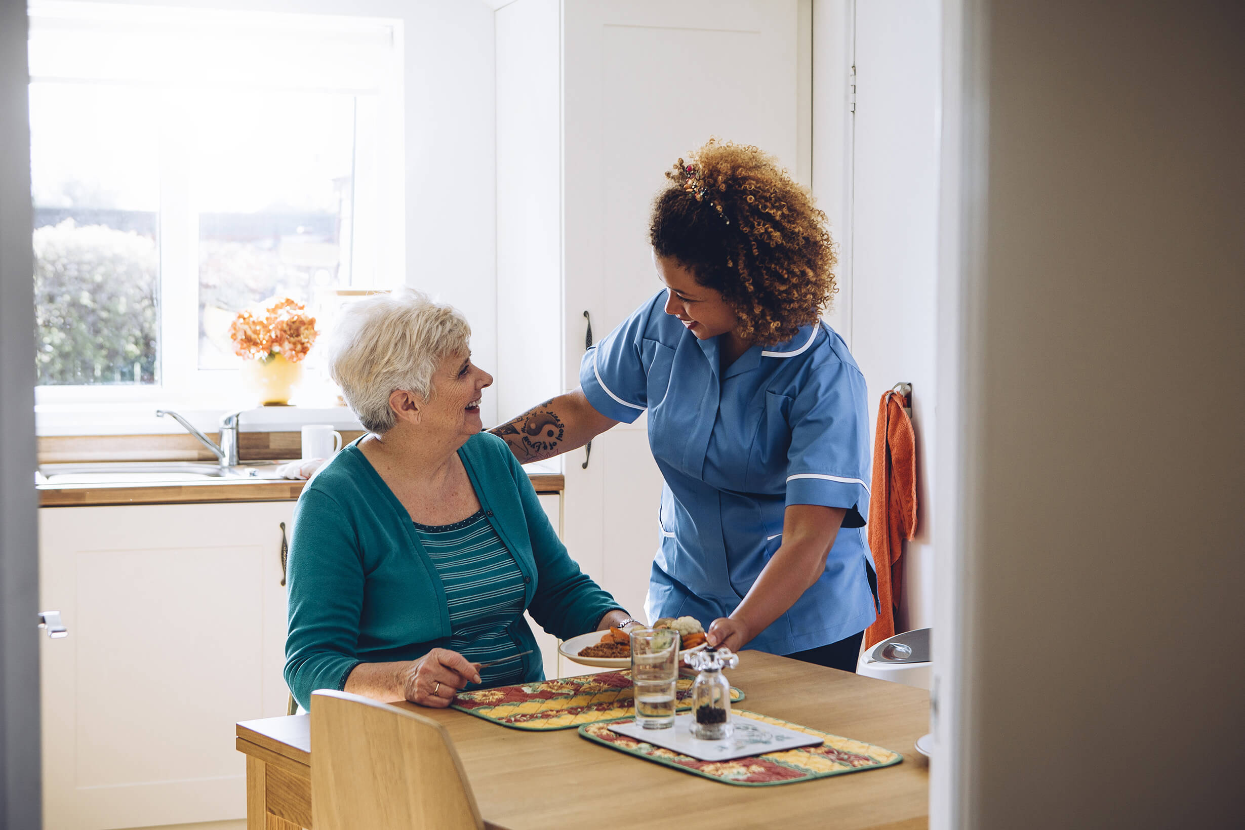 A nurse and an elderly patient at a table. The nurse is giving the lady her dinner.