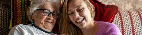 An eldery lady with glasses is sitting on a sofa laughing with a younger lady with long blonde hair, who is her personal assistant